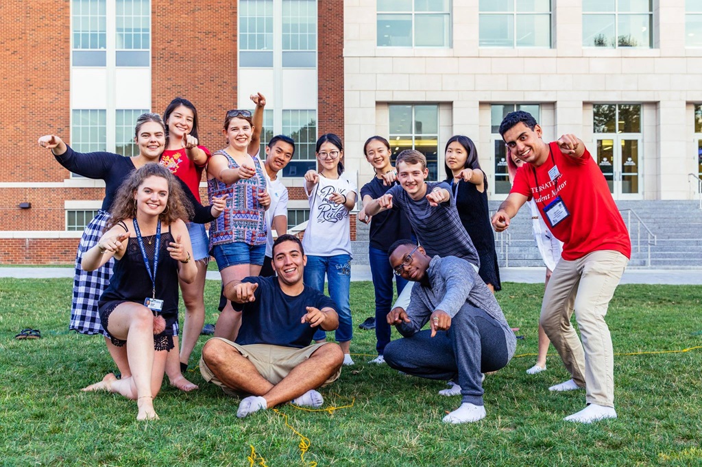 group of university students of all types on a university lawn pointing at the camera